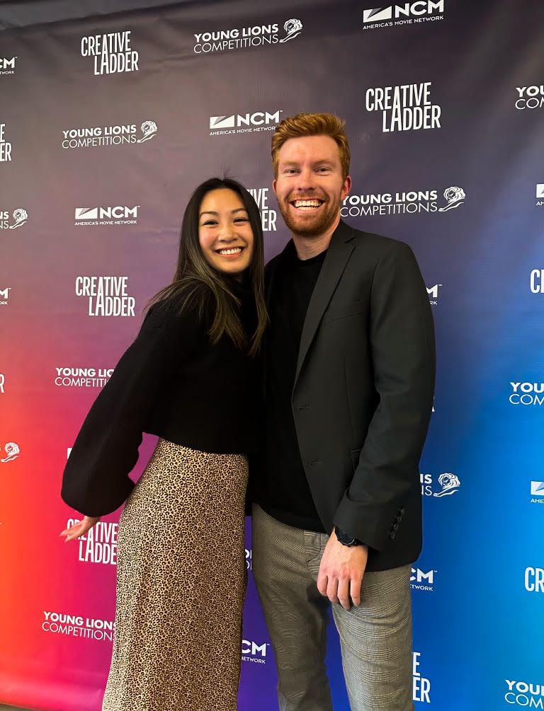Trevor Dunne, Associate Designer, a young man with red hair and a big smile stands with his arm around his partner, a young asian women wearing a black top and leopard print dress. He is wearing a grey sports coat and plaid pants and they are both standing in front of a colorful gradient of background with the logos of National CineMedia, Young Lions Competition and Creative Ladder. On top of this image is copy that reads, "3 Tips for Young Creatives from a Young Lions Finalist."