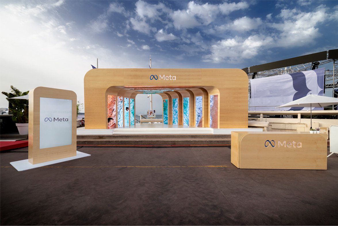 A large, wooden, organic shaped entrance is erected outdoors before a beach. The wooden entrance has the Meta logo engraved into the top center. The entrance forms a tunnel to the beach with screens on both sides of the tunnel revealing different beach textures. Next to the entrance is a large screen that says Meta and a wooden table. 