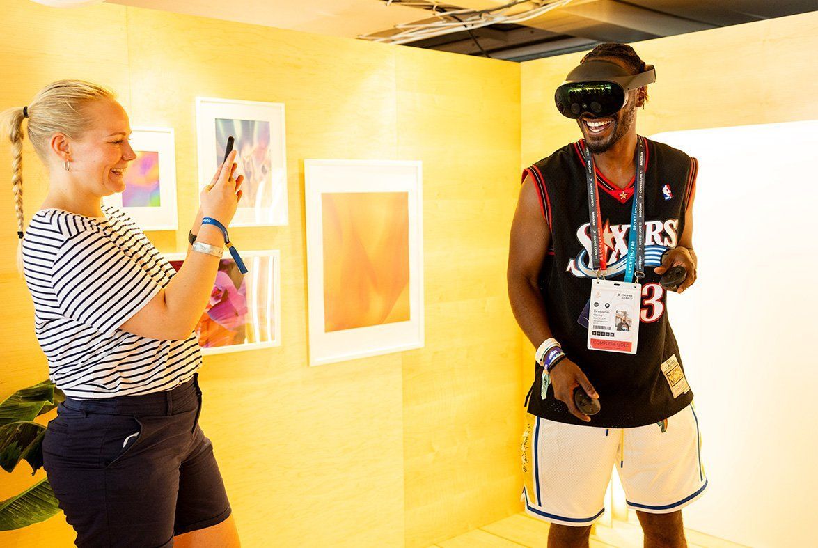 A person wearing virtual reality goggles and a person standing in front of a wall Description automatically generated 