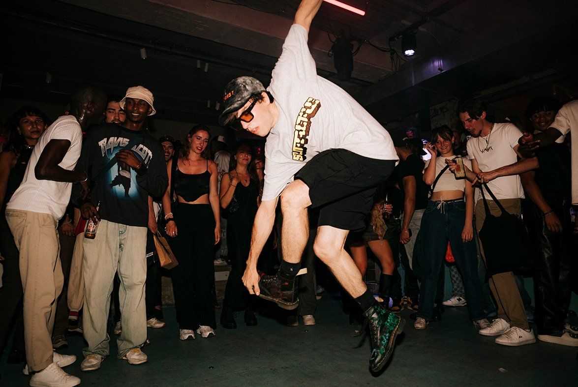 Man in a camo cap, orange glasses, grey t-shirt and black shorts doing a breakdancing move. He is holding one foot in the air and jumping off the ground wearing doc martin boots. He is surrounded by people cheering him on.