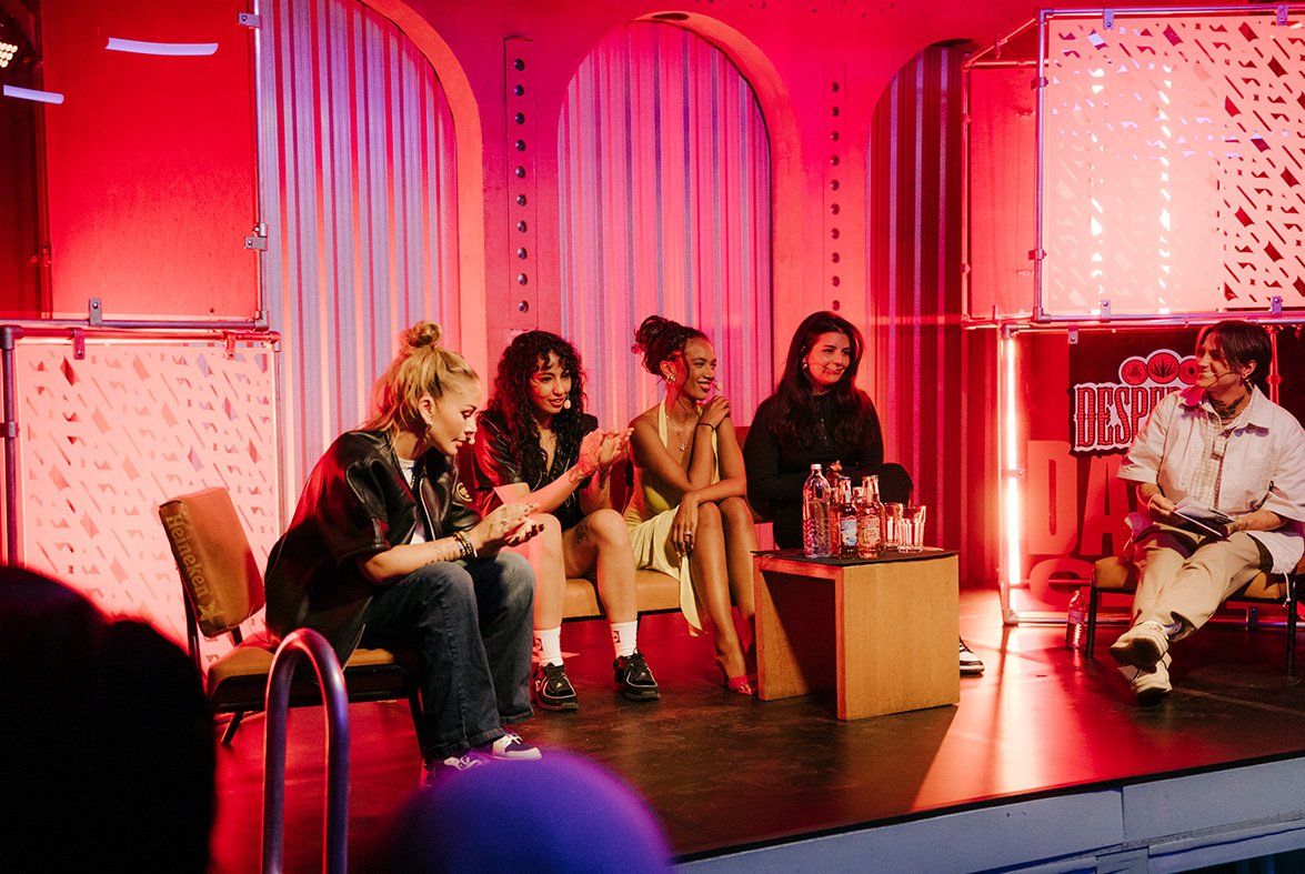 A group sitting on a stage having a panel discussion. On the left hand side are 4 females with microphones sitting and smiling, talking to each other and looking out off the stage. In front of them is a square table with drinks and Desperados bottles. To the right of them is a fifth person with a microphone and que cards. Behind them is a red lit wall with Desperados branded cube structures.