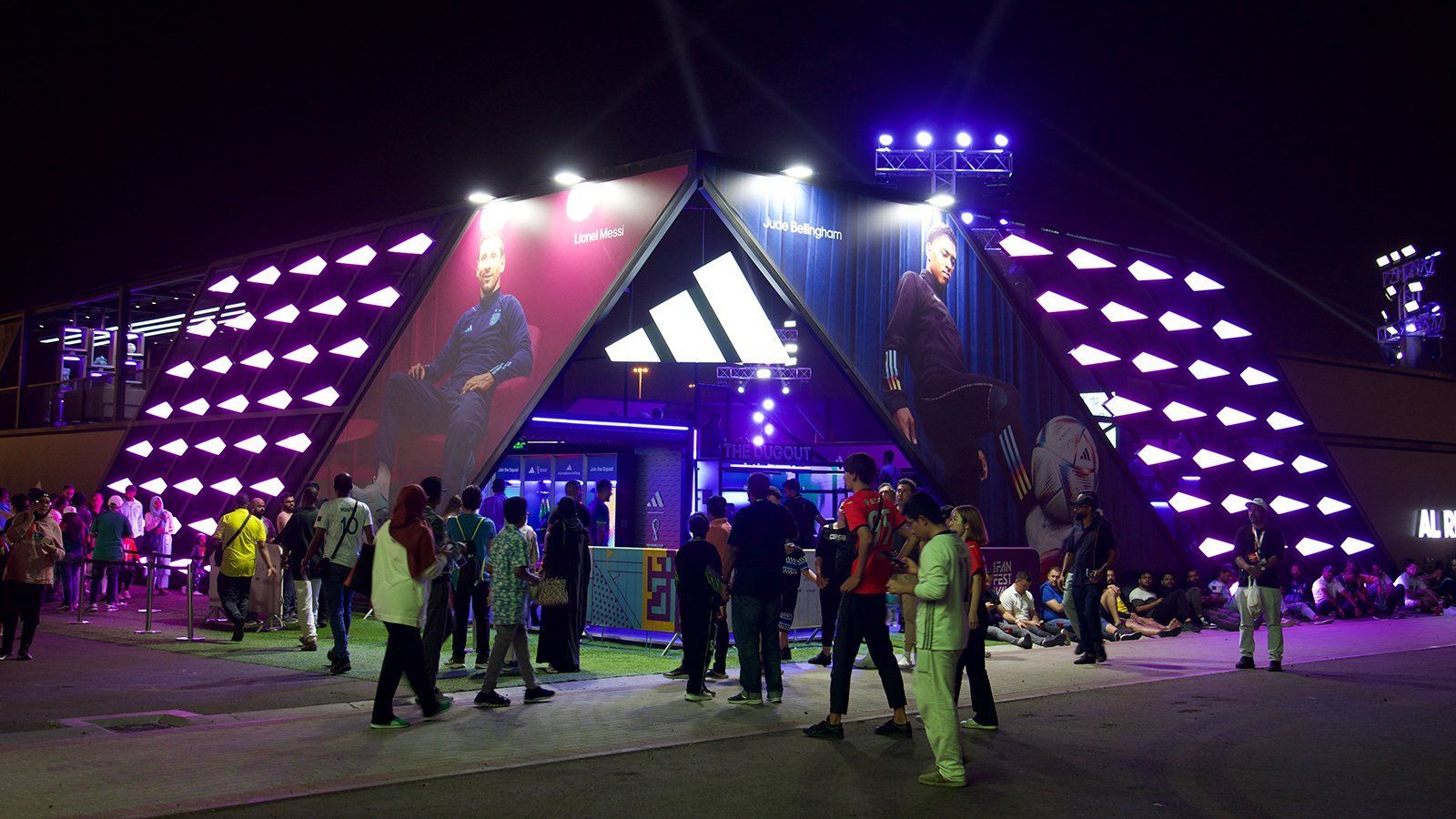 A large structure is erected from the ground with the adidas logo brightly illuminated at the top. It is dark out and vibrant blue and red color tones project from the structure and the entrance where crowds of people are walking in.