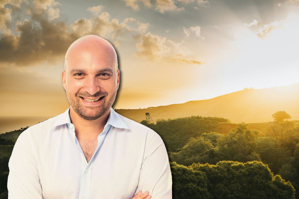 Julien Le Bas, a smiling man with a shaved head and short goatee, wearing a white button down shirt, set against a forest canopy sunset backdrop